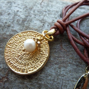 Könnte beinhalten: Eine goldene Halskette mit einem runden, gravierten Anhänger mit einem kreisförmigen Design und einer kleinen weißen Perle. Die Halskette besteht aus einem braunen Lederband.