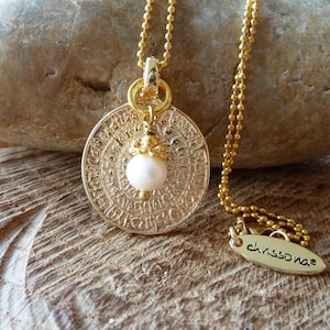 Könnte beinhalten: Goldkette mit einem runden Anhänger mit filigranen Details und einer weißen Perle. Der Anhänger ist mit Text beschriftet. Die Kette liegt auf einer Holzoberfläche mit einem Stein und einem Etikett mit der Aufschrift "chrissonae".