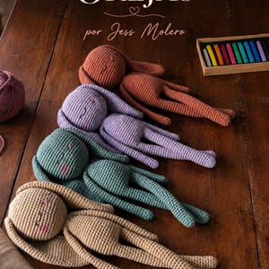 Puede incluir: Cuatro muñecos de conejo tejidos a ganchillo hechos a mano con orejas largas y caras bordadas, dispuestos en fila. Los muñecos son de color óxido, lavanda, verde azulado y beige. El texto OREJAS por Jess Molero es visible arriba.