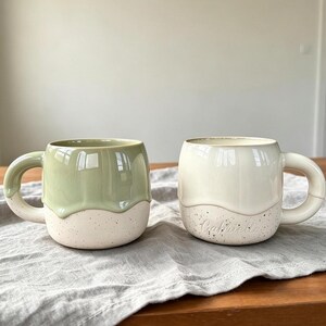 May include: Two ceramic mugs with rounded handles. The left mug features a sage green glazed top half and a speckled cream base. The right mug is entirely cream-colored with a speckled base and the name Gabriella embossed on the lower side.