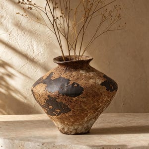 May include: A wide-mouthed ceramic vase with a textured, mottled surface featuring patches of dark charcoal black and earthy brown tones. The vessel has a rounded, tapered body and contains a cluster of thin, dried, light brown botanical stems.