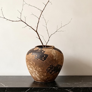 May include: A round, bulbous ceramic vase with a textured, mottled surface featuring patches of dark brown and earthy tan tones. The vase has a wide, open mouth and contains a single, thin, bare branch extending upward from the center opening.