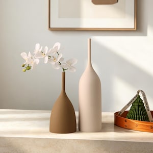 May include: Two tall, slender vases with matte finishes, one in dark brown and one in light beige. The brown vase holds a white orchid stem. Beside them sits a green, pleated, cone-shaped object inside a circular orange tray.