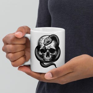 May include: A white ceramic mug featuring a detailed black and white graphic illustration of a human skull with a snake coiled around it. The snake's head rests on the top of the cracked skull, with its tongue extended, creating a high-contrast design.
