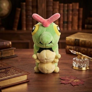 Pode incluir: Um peluche em forma de lagarta verde com a barriga amarelo-claro, olhos grandes amarelos e uma antena rosa em forma de V na cabeça. A criatura macia e recheada está de pé, com um corpo segmentado e pequenos membros.