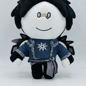 Puede incluir: Un muñeco de peluche con cabello negro, cara blanca y ojos y boca bordados. Viste una túnica azul con un emblema solar blanco en el pecho, una faja gris y pantalones negros. Estructuras oscuras en forma de alas sobresalen detrás de los hombros.