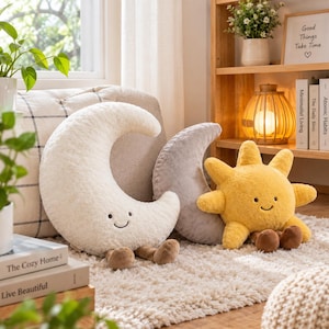 Peut inclure: Un ensemble de trois coussins en peluche comprenant un grand croissant de lune crème, un croissant de lune gris plus petit et un soleil jaune vif. Chaque coussin présente un visage souriant brodé et de petites jambes et pieds bruns.