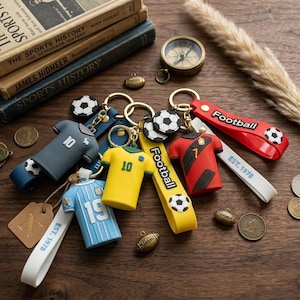 May include: A collection of sports-themed keychains featuring miniature soccer jerseys in blue, yellow, and red, along with white straps labeled Football and EST. 1978. The set includes small soccer ball charms and bronze-toned football pendants.