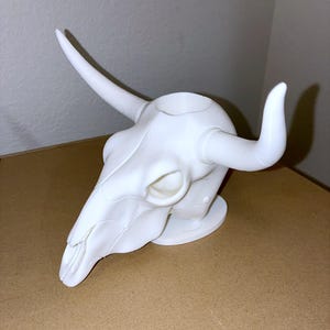 May include: A matte white 3D printed bull skull sculpture featuring long, curved horns and a hollow circular opening at the top of the cranium. The skull is mounted on a flat, oval-shaped base, showcasing a smooth surface with visible structural lines.