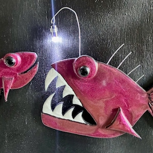 May include: Two decorative fish, one small and one large, in shades of pink with black eyes. The larger fish has a wide open mouth with white and black teeth and a small light above it. The background is black.