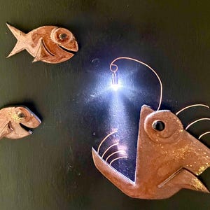 May include: Metal wall art featuring three fish. Two smaller fish flank a larger anglerfish with an open mouth and a glowing light. The fish are bronze-colored with gold accents, set against a dark background.