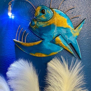 May include: A blue and gold anglerfish wall art piece with a small light. The fish has a detailed design with a light blue body and gold accents. The art is mounted on a blue wall, with white decorative feathers at the bottom.