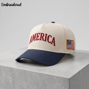 Könnte beinhalten: Eine cremefarbene Baseballkappe mit marineblauem Schirm. Auf der Vorderseite ist das Wort AMERICA in roten Großbuchstaben eingestickt. Ein kleiner rechteckiger Aufnäher der Flagge der Vereinigten Staaten ist an der Seite der Kappe angebracht.