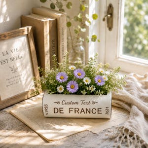 May include: A rectangular white planter shaped like a book, featuring the text Custom Text DE FRANCE N 1955 2631. The planter is filled with a floral arrangement of purple daisies, small white flowers, and green foliage, creating a decorative display.