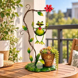 May include: A metal garden ornament featuring a green frog sitting on a swing suspended from a brown vine-like structure with green leaves and a red flower. A copper-coloured planter with the word Bloom and a bee graphic holds succulents.