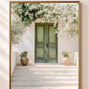 May include: A framed wall art print depicting a set of double doors painted in a muted green, set into a white wall. The entrance is framed by white flowering vines and features stone steps leading up to the doors with two terracotta pots.