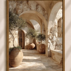 Op de afbeelding: Een ingelijste wandkunstprint met een stenen boogcorridor met beige getextureerde muren en een lichtgekleurde stenen vloer. Drie grote terracotta-kleurige keramische urnen met kleine groene olijfbomen staan opgesteld langs het looppad.