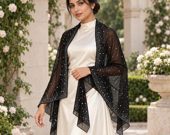 May include: A sheer black shawl featuring a dense pattern of silver rhinestones along the edges and scattered across the fabric, worn over a sleeveless cream-colored satin dress with a high neckline and a smooth, flowing silhouette.