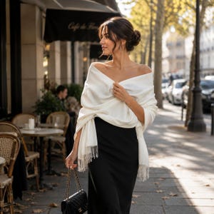 May include: A soft white pashmina shawl with delicate fringe edges draped over the shoulders, paired with a simple black dress and a small black quilted handbag featuring a gold chain strap and a gold logo emblem on the front flap.