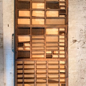 May include: A rectangular wooden printer's type drawer featuring numerous small compartments of varying sizes. The interior base of each section is lined with light tan paper. A metal handle is attached to the left side of the frame.