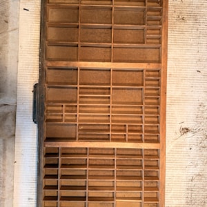 May include: A rectangular wooden printer's tray featuring a grid of numerous small, open compartments of varying sizes. The frame is constructed from medium-toned wood, and the interior dividers create a structured, multi-sectioned storage layout.