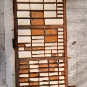 May include: A rectangular wooden printer's type case featuring a grid of numerous small, open compartments of varying sizes. The frame is constructed from dark brown wood, while the interior dividers are made of lighter wood with white paper inserts.