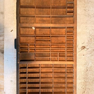 May include: A rectangular wooden printer's drawer featuring a grid of numerous small, open compartments of varying sizes. The wood has a natural brown finish with visible grain and a dark edge along the left side, designed for storage.