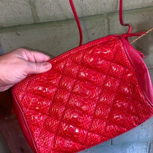 May include: A vibrant red crossbody bag featuring a quilted diamond pattern with a textured snakeskin-like finish. The bag includes a thin matching red shoulder strap and a smooth red leather side panel, providing a structured rectangular shape.