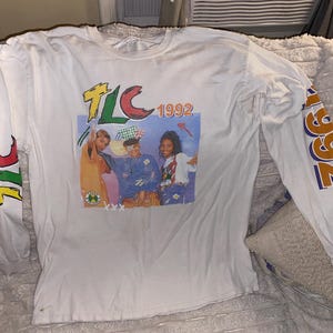 May include: A white long-sleeve t-shirt featuring a graphic print of the music group TLC with the text TLC 1992. The sleeves are printed with the text TLC and 1992 in yellow, red, and blue. The graphic shows three individuals in colourful clothing.