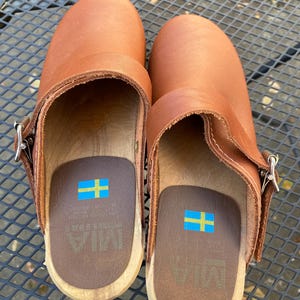 May include: A pair of tan leather clogs with wooden soles and silver-toned metal buckles on the sides. The interior footbeds are brown with a printed Swedish flag and the brand name MIA visible in the center of the heel area.