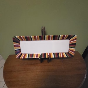 May include: A rectangular serving platter featuring a solid white center and a wide border decorated with vertical stripes in shades of red, yellow, green, black, and white. The platter is held upright by a dark brown wooden display stand.