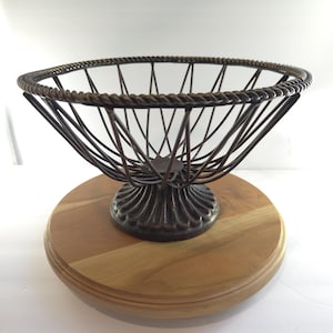 May include: A dark bronze-colored wire basket with a twisted rope-style rim and a fluted pedestal base. The basket is mounted on a circular, light-toned wooden base with a smooth, natural grain finish and a beveled edge detail.