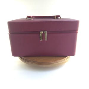 May include: A rectangular, deep magenta cosmetic train case with a textured surface. The case features a top handle, a zippered closure with two gold-toned metal pulls, and the brand name Estée Lauder engraved on the zipper pulls.