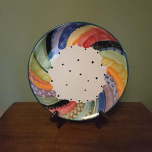 Puede incluir: Un plato redondo de cerámica con un centro blanco decorado con pequeños puntos negros. El borde está adornado con una vibrante cenefa multicolor de formas curvas y abstractas en tonos azul, naranja, verde, rojo y morado con patrones.