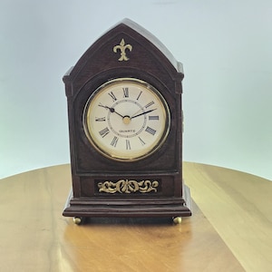 May include: A dark brown wooden mantel clock with a pointed gothic-style arch top. The clock face features a cream background, black Roman numerals, and the word QUARTZ. A gold-colored fleur-de-lis emblem sits above the dial, with gold filigree trim.