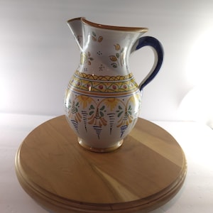 May include: A ceramic pitcher with a white base featuring hand-painted yellow, green, and blue floral and geometric patterns. The vessel has a curved handle, a wide spout, and a rounded body, showcasing intricate decorative motifs around the exterior.