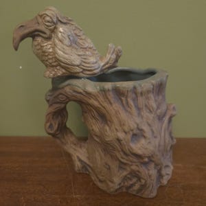 May include: A ceramic mug shaped like a textured tree stump with a sculpted bird perched on the rim. The mug features a brown, wood-grain finish with a handle integrated into the side. The interior of the mug is a smooth, light blue-grey color.