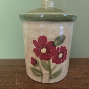 May include: A cylindrical ceramic canister with a removable lid featuring a knob handle. The canister has a cream-colored base with a hand-painted design of two large dark red flowers with yellow centers and green leaves on the front surface.