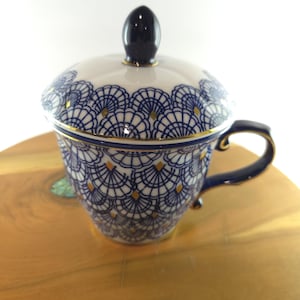 May include: A white porcelain teacup with a lid, featuring a repeating blue fan-shaped pattern with small gold diamond accents. The cup has a dark blue handle and a matching dark blue finial on the lid, with gold trim along the rim and handle edges.