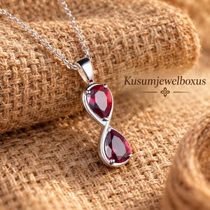 Puede incluir: Un collar de plata con un colgante de dos piedras preciosas de color rojo intenso en forma de pera, dispuestas verticalmente en forma de infinito. El engaste es de plata pulida. El texto Kusumjewelboxus es visible a la derecha.