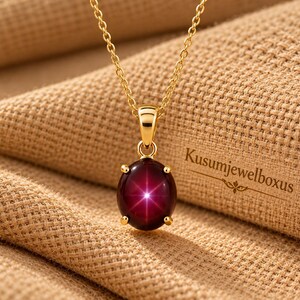 May include: A gold chain necklace featuring a round, deep red cabochon pendant with a prominent six-rayed star effect centred on the stone. The pendant is held by four gold prongs. The text Kusumjewelboxus is visible to the right of the pendant.