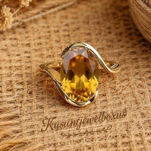 Può includere: Un anello in oro con una grande pietra preziosa citrino gialla a taglio ovale come elemento centrale. La fascia ha una finitura in oro lucido, liscia e curva che avvolge la pietra. Il testo Kusumjewelboxus è stampato in corsivo sotto l'anello.