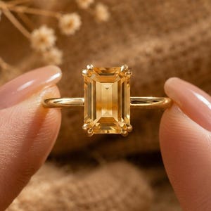 May include: A gold ring featuring a rectangular emerald-cut yellow gemstone set in a four-prong setting. The band is thin and polished gold. The text Kusumjewelboxus is visible in the upper right corner of the image.
