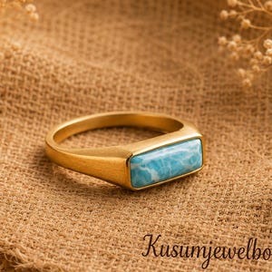 May include: A gold-toned ring featuring a rectangular, light blue, marbled stone inlay. The band has a smooth, polished finish. The text 'Kusumjewelboxus' is printed in a dark script font below the ring, accompanied by a small heart icon.