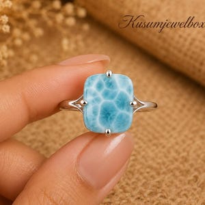 May include: A silver ring featuring a rectangular, light blue Larimar gemstone with a distinct white marbled pattern. The stone is held in a four-prong setting on a thin, polished silver band. The text Kusumjewelboxus is visible in the corner.