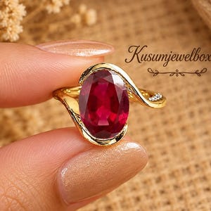May include: A gold ring featuring a large, oval-cut, deep red gemstone set in a bypass-style band. The band includes small, clear accent stones on one side. The text Kusumjewelboxus is visible in the upper right corner of the image.