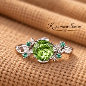 May include: A silver-toned ring featuring a large, round, faceted lime-green centre stone flanked by smaller, round, dark-green accent stones. The band is designed with intricate leaf-like metalwork. The text Kusumjewelboxus is visible.