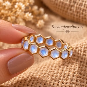 May include: A gold-toned ring featuring a series of connected hexagonal settings, each containing a small, iridescent blue moonstone. The text 'Kusumjewelboxus' is visible in the upper right corner, accompanied by a small lotus flower icon.