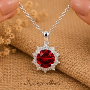 May include: A silver necklace featuring a round, deep red gemstone pendant surrounded by a halo of small, clear, sparkling stones in a starburst setting. The pendant hangs from a delicate silver chain, with the text Kusumjewelboxus below.