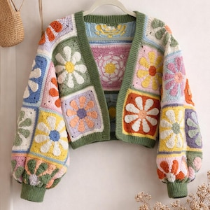 May include: A colorful, crocheted cardigan with a floral patchwork design. The open-front sweater features square panels in various colors, including white, yellow, orange, pink, and blue, with green trim and cuffs. The sleeves are puffy.